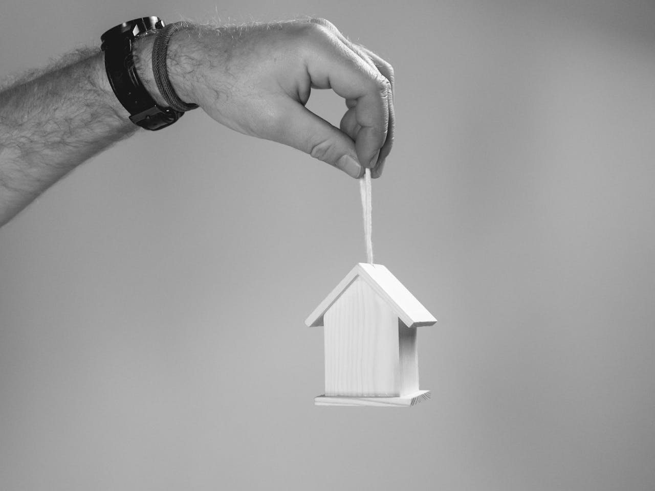Home Black and white photo of a hand holding a miniature wooden house by a string, symbolizing real estate.